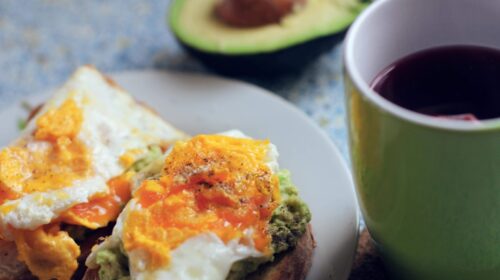 plate of breakfast sandwich beside avocado and hot beverage